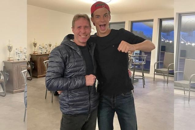 Zwei junge M&auml;nner umarmen sich, l&auml;chelnd vor einem Raum mit Pokalen und St&uuml;hlen im Hintergrund.