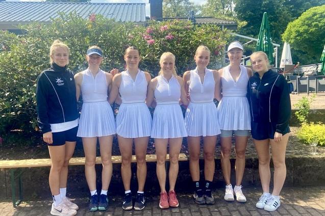 Gruppe von sieben Tennis-Spielerinnen in wei&szlig;en Dresses, l&auml;chelnd vor einer gr&uuml;nen Kulisse im Freien.