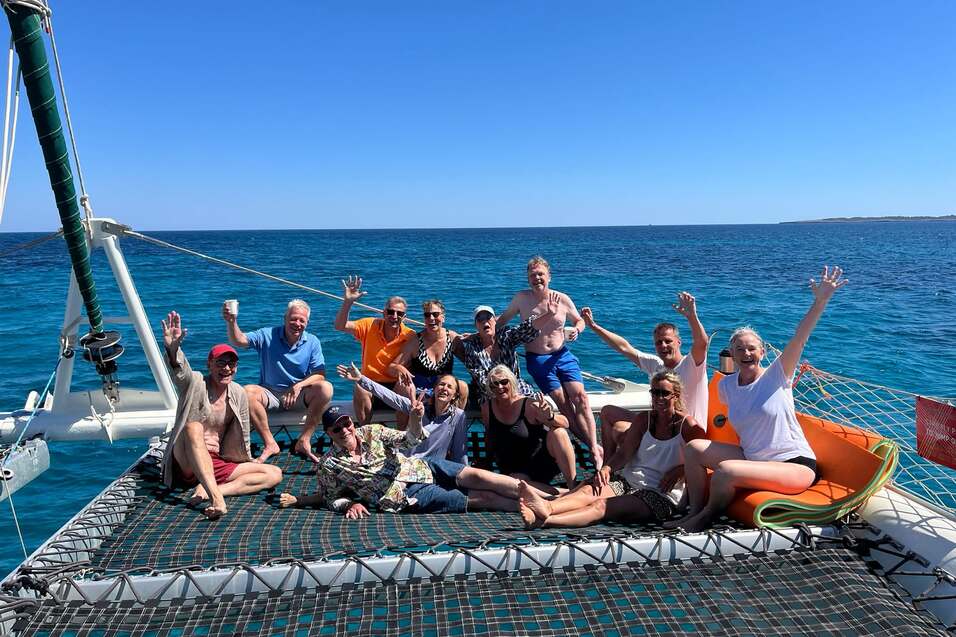 Gruppe von 15 Personen auf einem Katamaran-Netz, l&auml;chelnd und winkend, mit blauem Meer im Hintergrund.