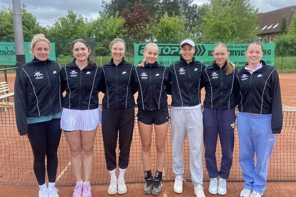 Sieben Tennisspielerinnen in Sportjacken stehen l&auml;chelnd auf einem Sandplatz vor einem Netz.