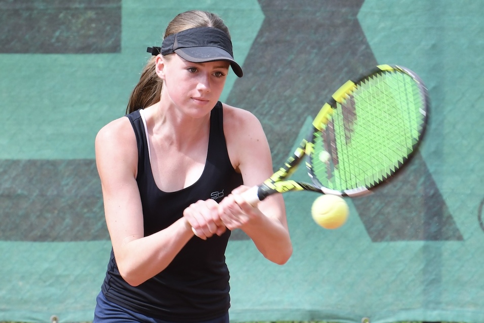 Junge Frau schl&auml;gt mit einem Tennisschl&auml;ger einen gelben Tennisball auf einem gr&uuml;nen Tennisplatz.