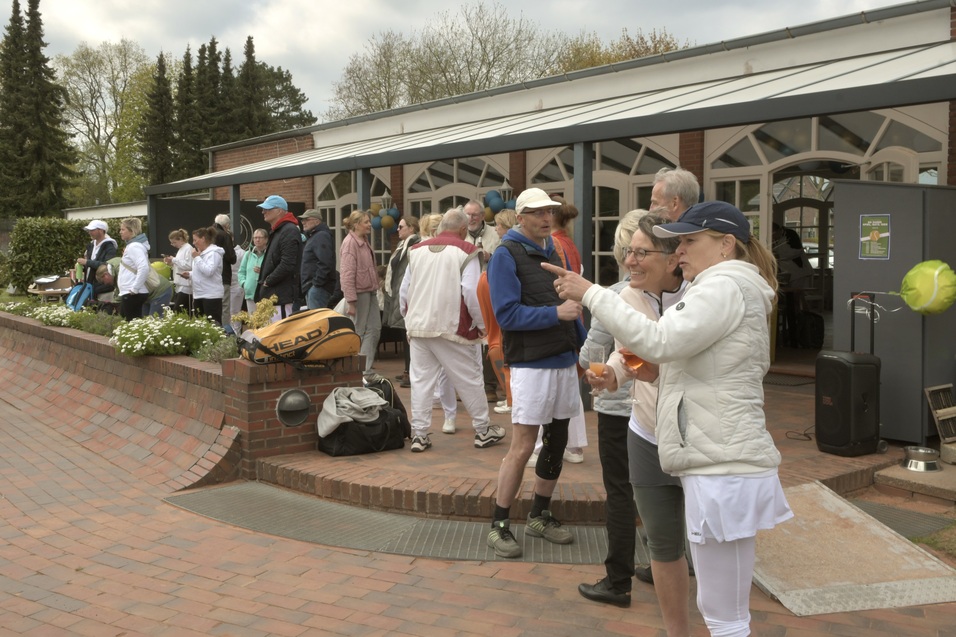 Menschen in Sportkleidung stehen vor einem Tennisplatz, einige unterhalten sich, andere warten in geselliger Runde.