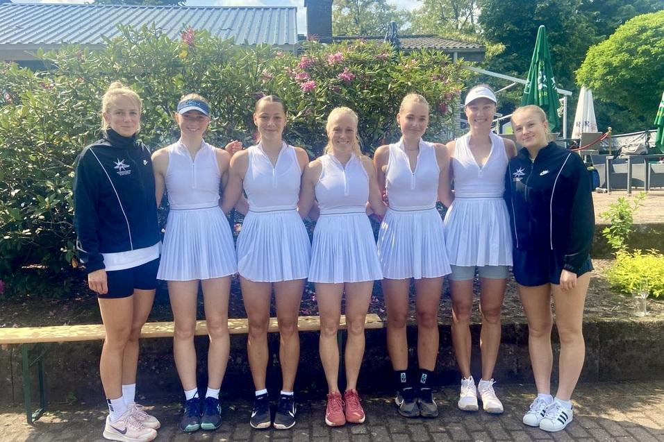 Gruppe von sieben Tennis-Spielerinnen in wei&szlig;en Dresses, l&auml;chelnd vor einer gr&uuml;nen Kulisse im Freien.