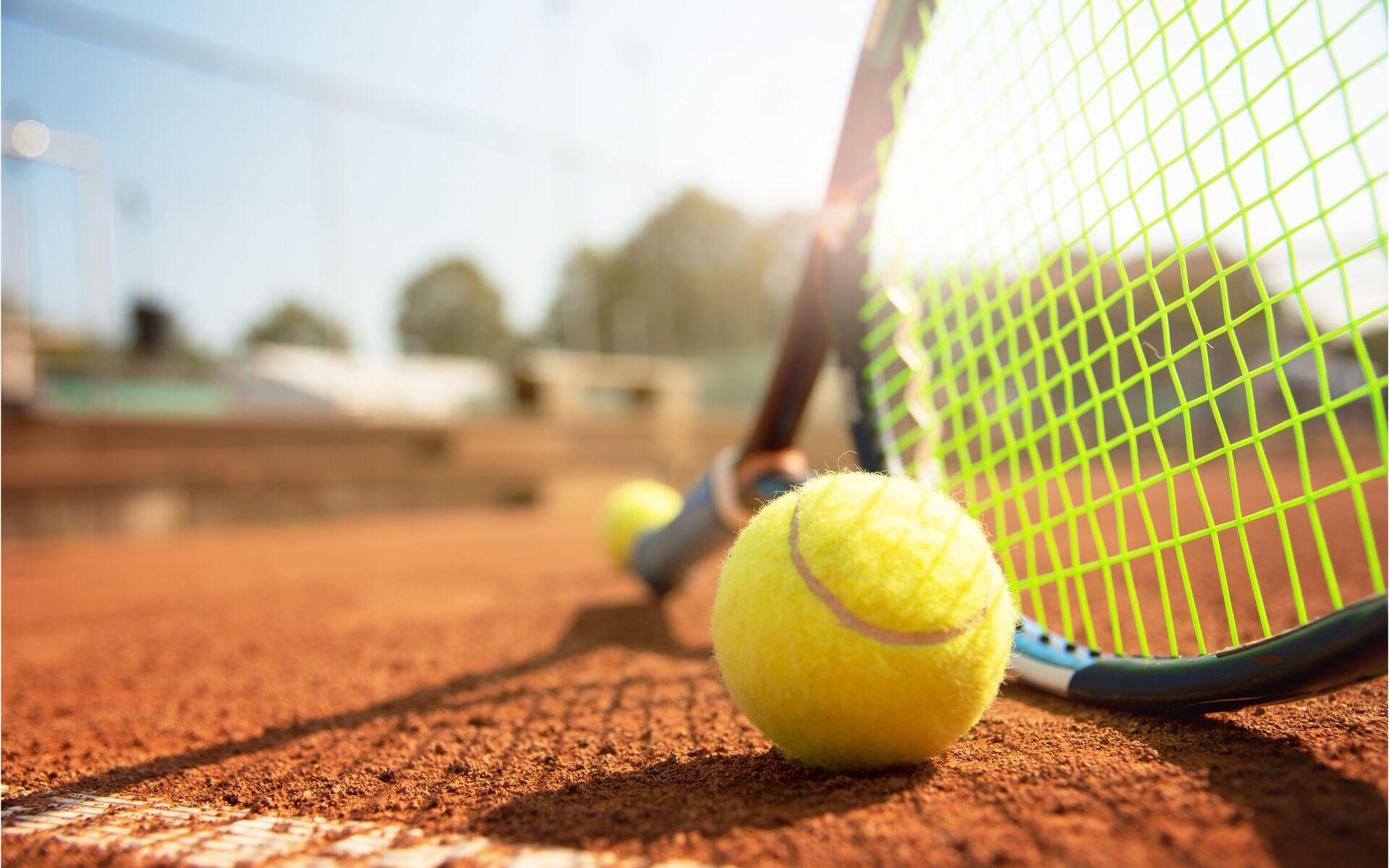 Nahaufnahme eines tennisballs auf einem roten Sandplatz, neben einem Tennisnetz und Schläger.