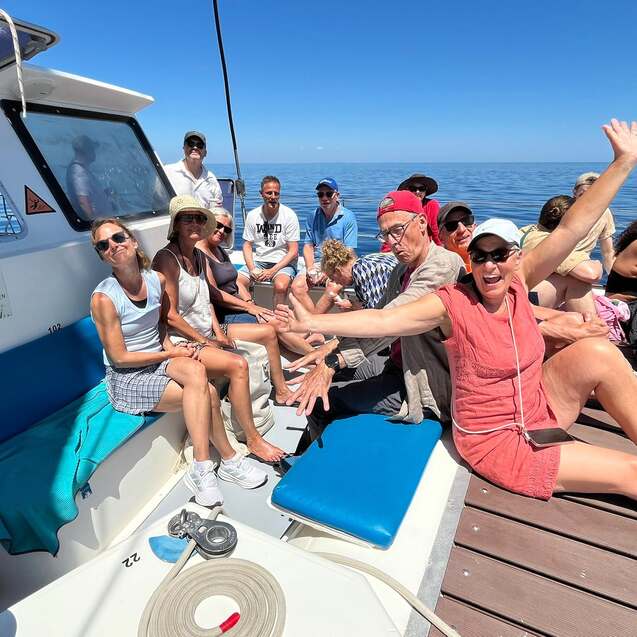 Gruppe von Personen entspannt auf einem Boot, einige winken, w&auml;hrend andere sich unterhalten, bei sonnigem Wetter.