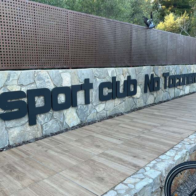 Schild des Sportclubs &bdquo;Na Tonnara&ldquo; an einer Steinmauer, umgeben von gr&uuml;ner Natur und Holzweg.