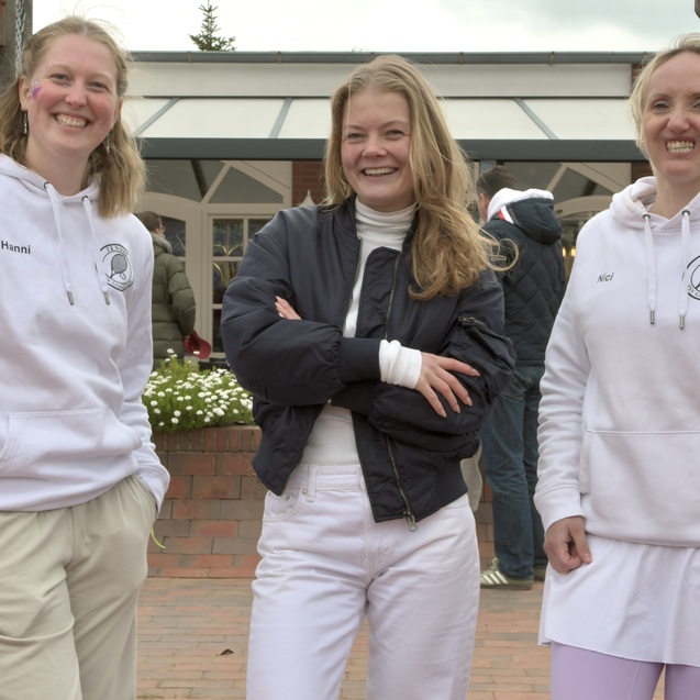 Drei Frauen in Sportkleidung stehen l&auml;chelnd vor einem Tennisplatz mit einem Zaun im Hintergrund.
