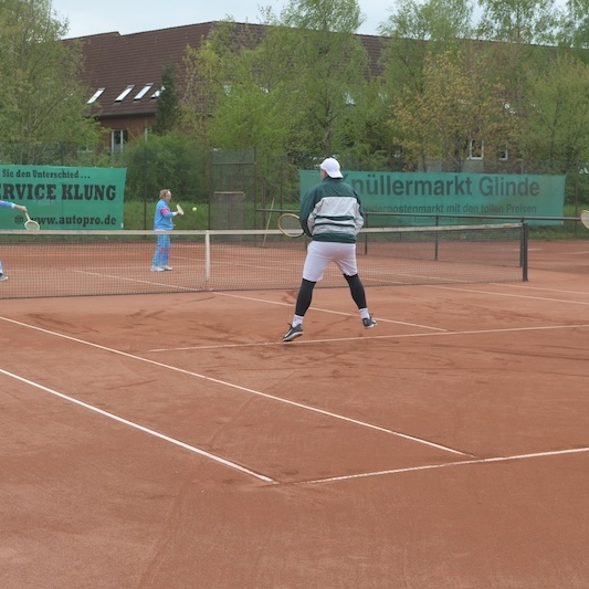 Drei Tennisspieler auf einem sandigen Platz, zwei in blauen Outfits, einer in grauer Jacke und schwarzer Hose, spielen ein Match.