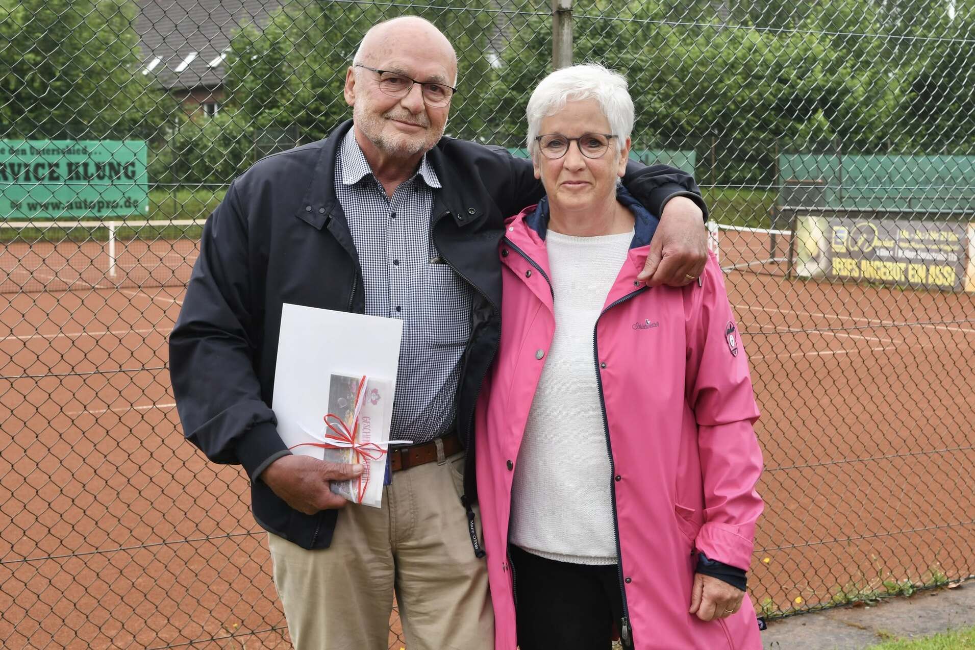 Ein &auml;lterer Mann und eine Frau stehen arm in arm vor einem Tennisplatz, der im Hintergrund sichtbar ist.