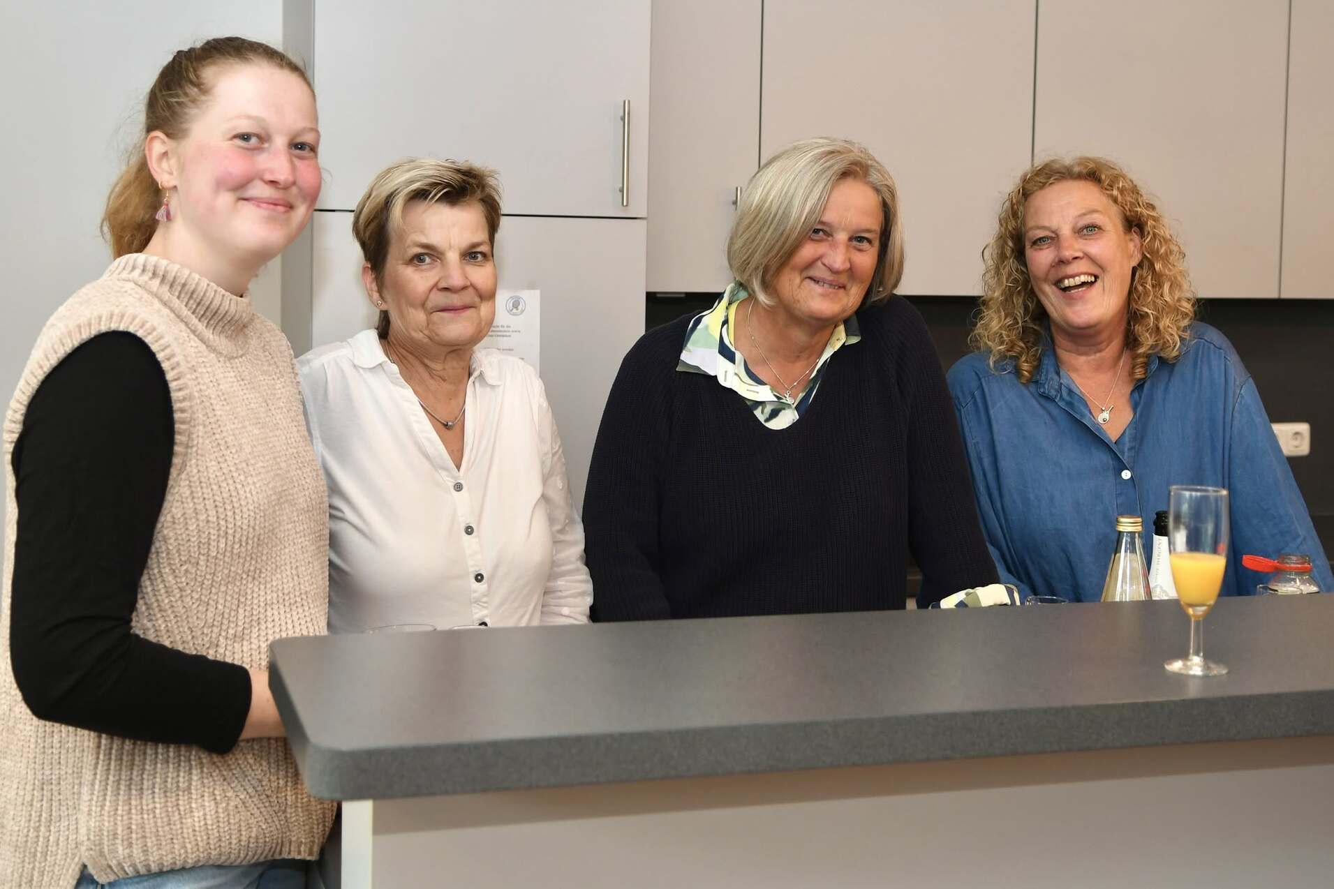 Vier Frauen stehen l&auml;chelnd an einer K&uuml;chenbar; eine h&auml;lt ein Glas mit Getr&auml;nk in der Hand.