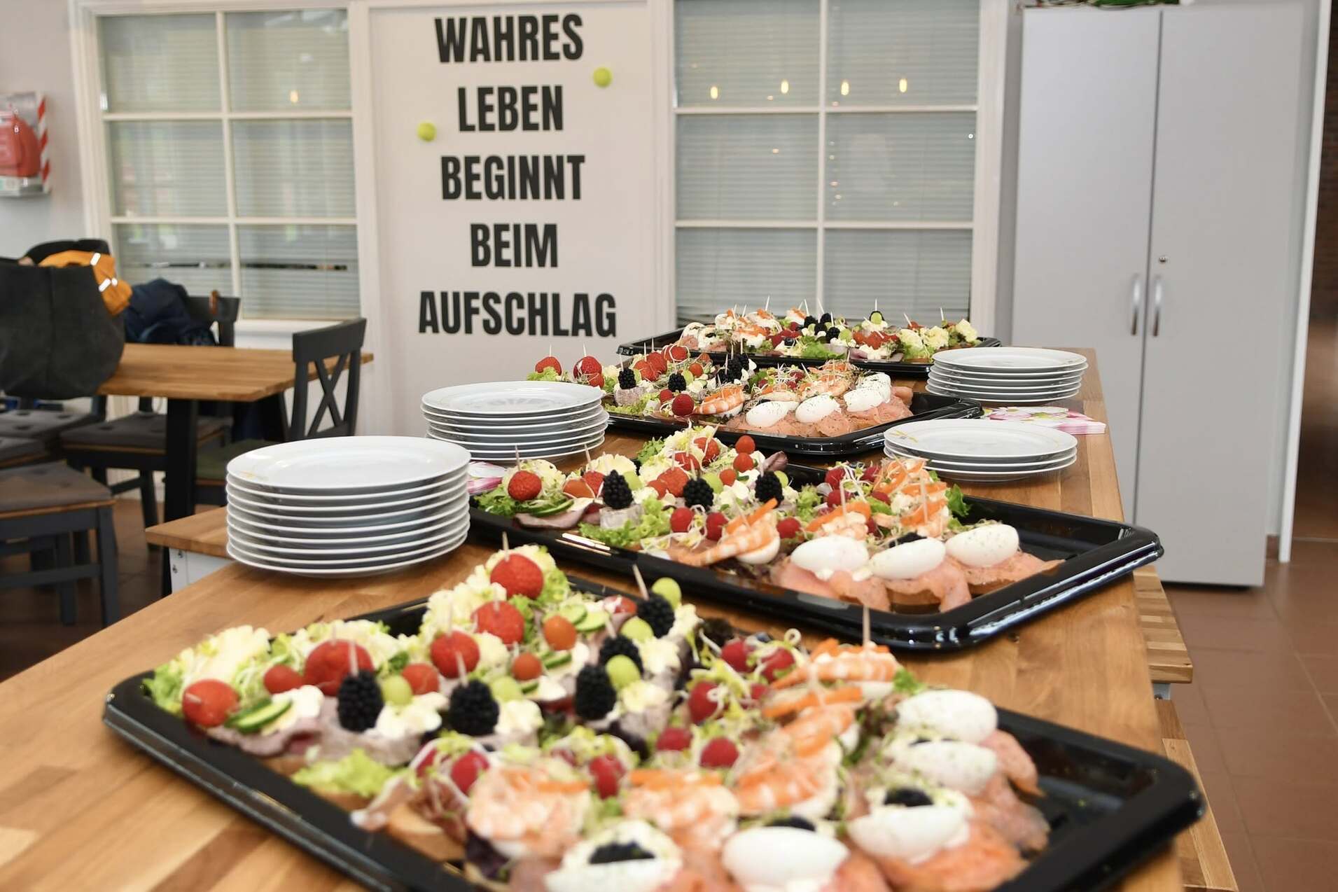 Buffet mit verschiedenen Vorspeisen, darunter Fisch und Gem&uuml;se, vor einer Wand mit dem Spruch &bdquo;WAHRES LEBEN BEGINNT BEIM AUFSCHLAG&ldquo;.