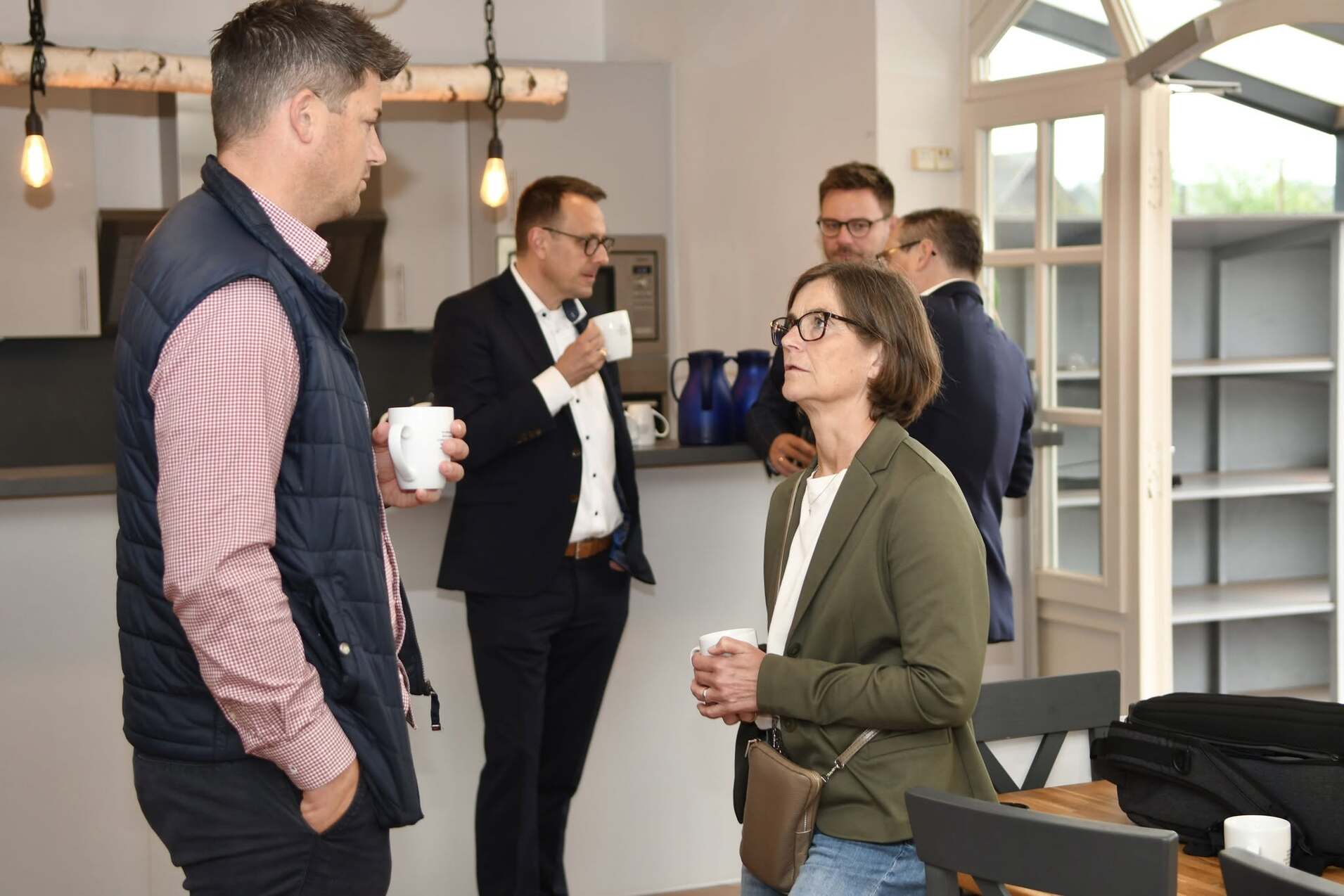 F&uuml;nf Personen stehen in einer hellen K&uuml;che und unterhalten sich bei Kaffeetassen in der Hand.