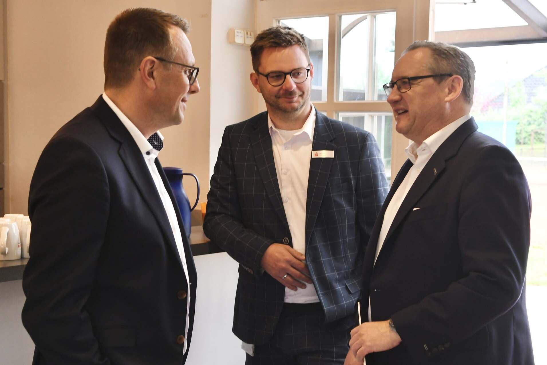 Drei M&auml;nner in Anz&uuml;gen stehen in einer hellen Umgebung und unterhalten sich freundlich.