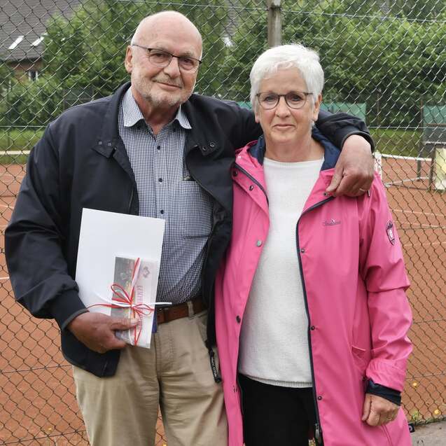 Ein &auml;lterer Mann und eine Frau stehen arm in arm vor einem Tennisplatz, der im Hintergrund sichtbar ist.