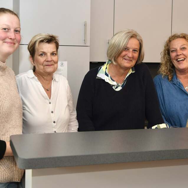Vier Frauen stehen l&auml;chelnd an einer K&uuml;chenbar; eine h&auml;lt ein Glas mit Getr&auml;nk in der Hand.