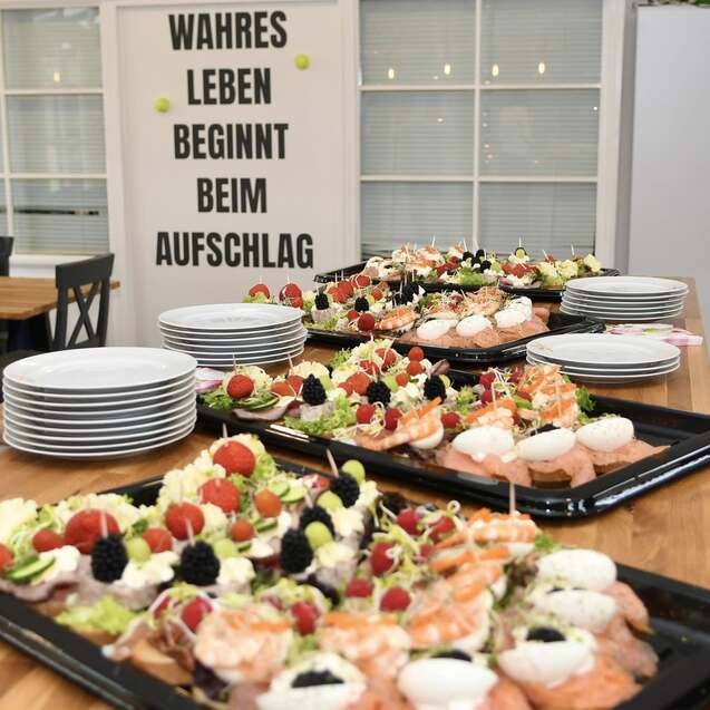 Buffet mit verschiedenen Vorspeisen, darunter Fisch und Gem&uuml;se, vor einer Wand mit dem Spruch &bdquo;WAHRES LEBEN BEGINNT BEIM AUFSCHLAG&ldquo;.