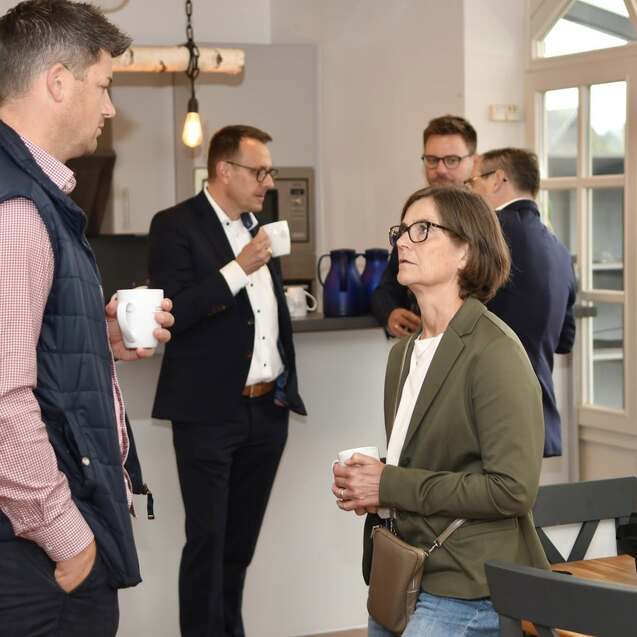 F&uuml;nf Personen stehen in einer hellen K&uuml;che und unterhalten sich bei Kaffeetassen in der Hand.
