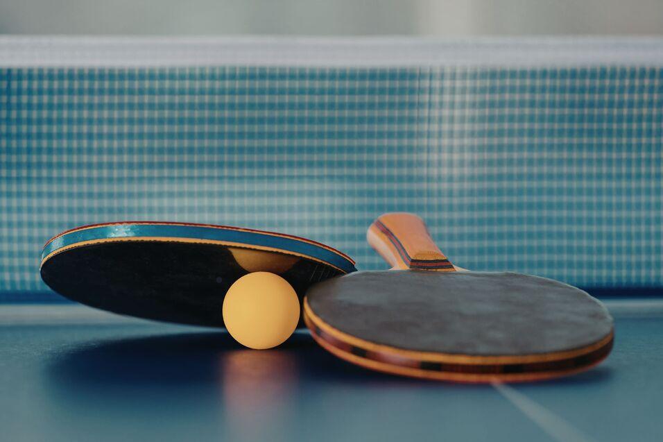 Nahaufnahme von zwei Tischtennisschlägern und einem gelben Ball auf einer blauen Tischtennisplatte.