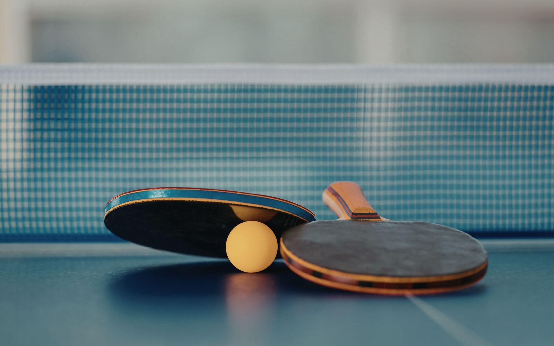 Nahaufnahme von zwei Tischtennisschlägern und einem gelben Ball auf einer blauen Tischtennisplatte.