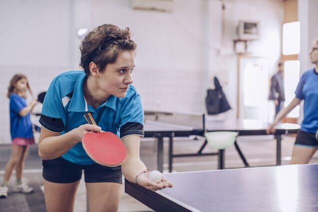 Junge Tischtennisspielerin mit Schläger und Ball, konzentriert an einem Tischtennistisch in einem Sportraum.