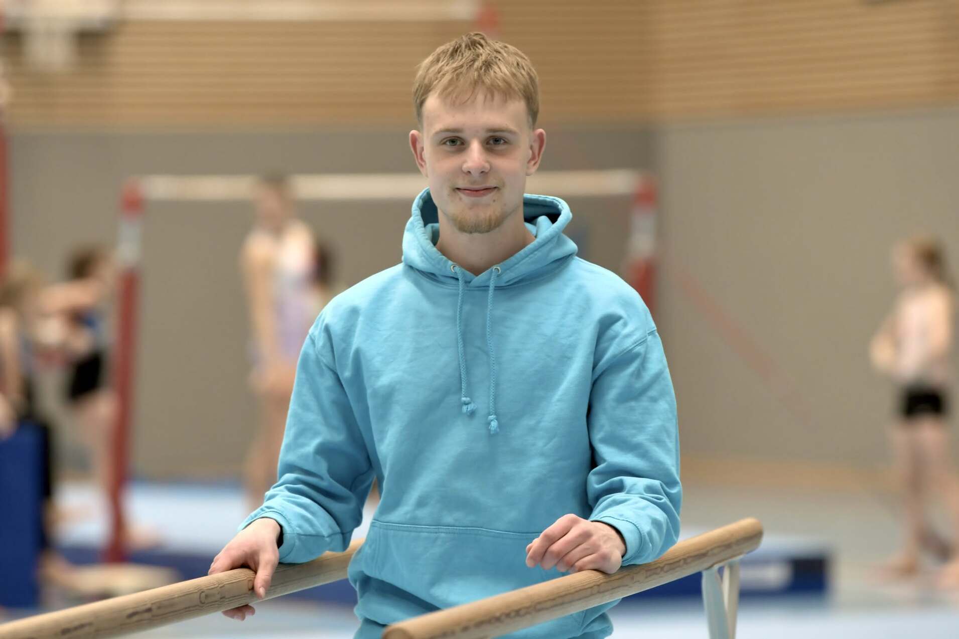 Junger Mann in blauer Kapuzenjacke steht an einem Barren in einer Sporthalle mit weiteren Turnern im Hintergrund.