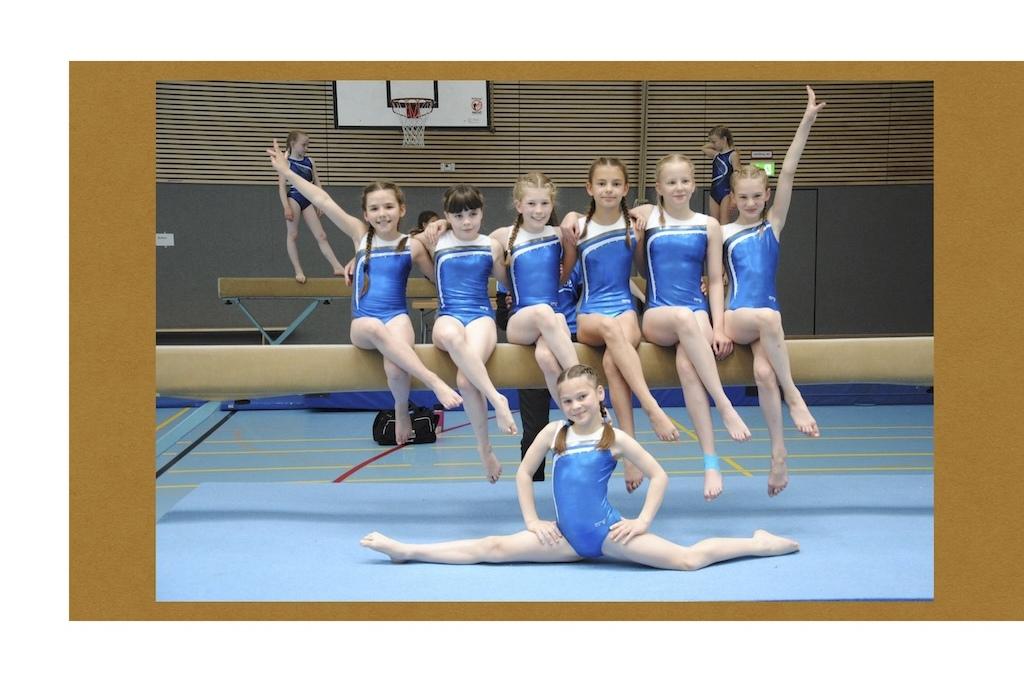 Gruppenfoto von acht jungen Turnerinnen in blauen Anzügen, die auf einem Schwebebalken sitzen und posieren.