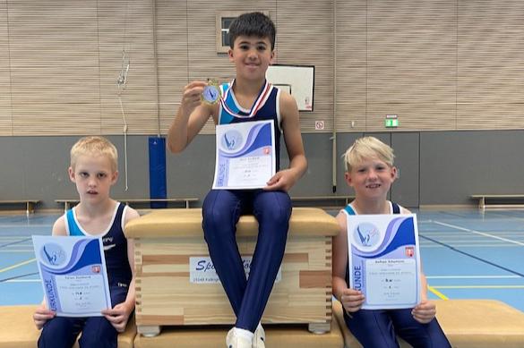 Drei Jungs sitzen auf einem K&uuml;rbis, halten Urkunden und eine Medaille, in einer sportlichen Halle.