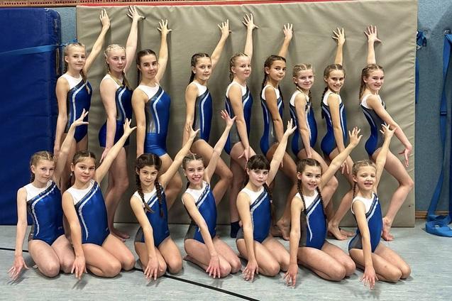 Gruppendfoto von 16 jungen Turnerinnen in blauen Wettkampfanz&uuml;gen, die in einer Sporthalle posieren.