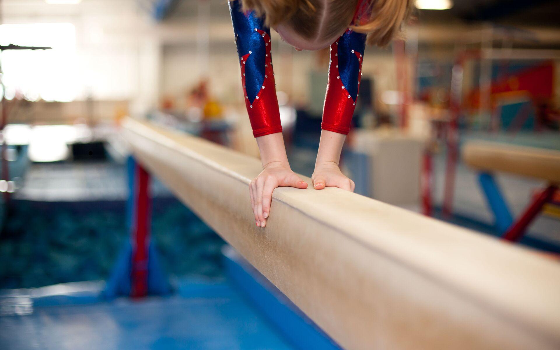 Mädchen in bodenlangem Anzug stützt sich auf einen Schwebebalken in einem Trainingsraum für Gymnastik.
