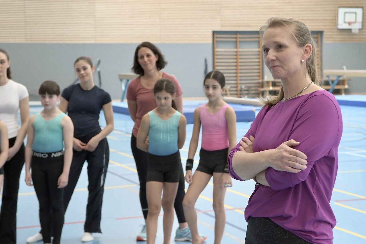 Gruppe junger Turnerinnen und Trainerinnen in einer Sporthalle, eine Trainerin beobachtet aufmerksam das Geschehen.