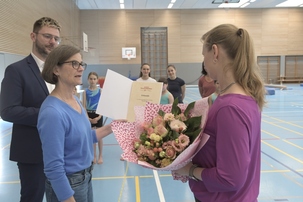 Frau &uuml;berreicht einer anderen Frau einen Blumenstrau&szlig; und ein Dokument in einer Sporthalle, im Hintergrund Sportlerinnen.