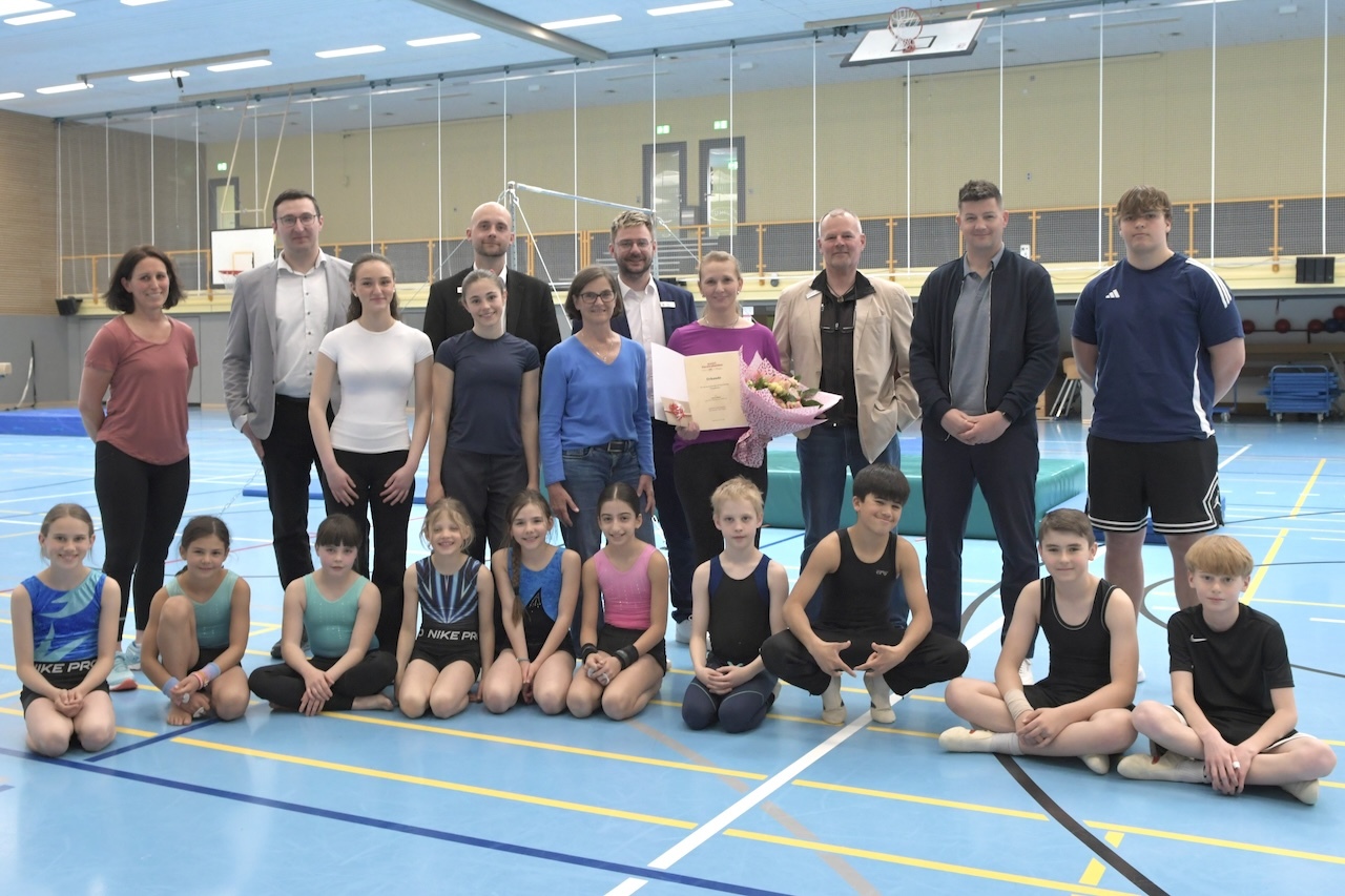 Gruppenausbildung mit jungen Turner*innen und Trainer*innen in einer Sporthalle, umgeben von Erwachsenen und Urkunden.