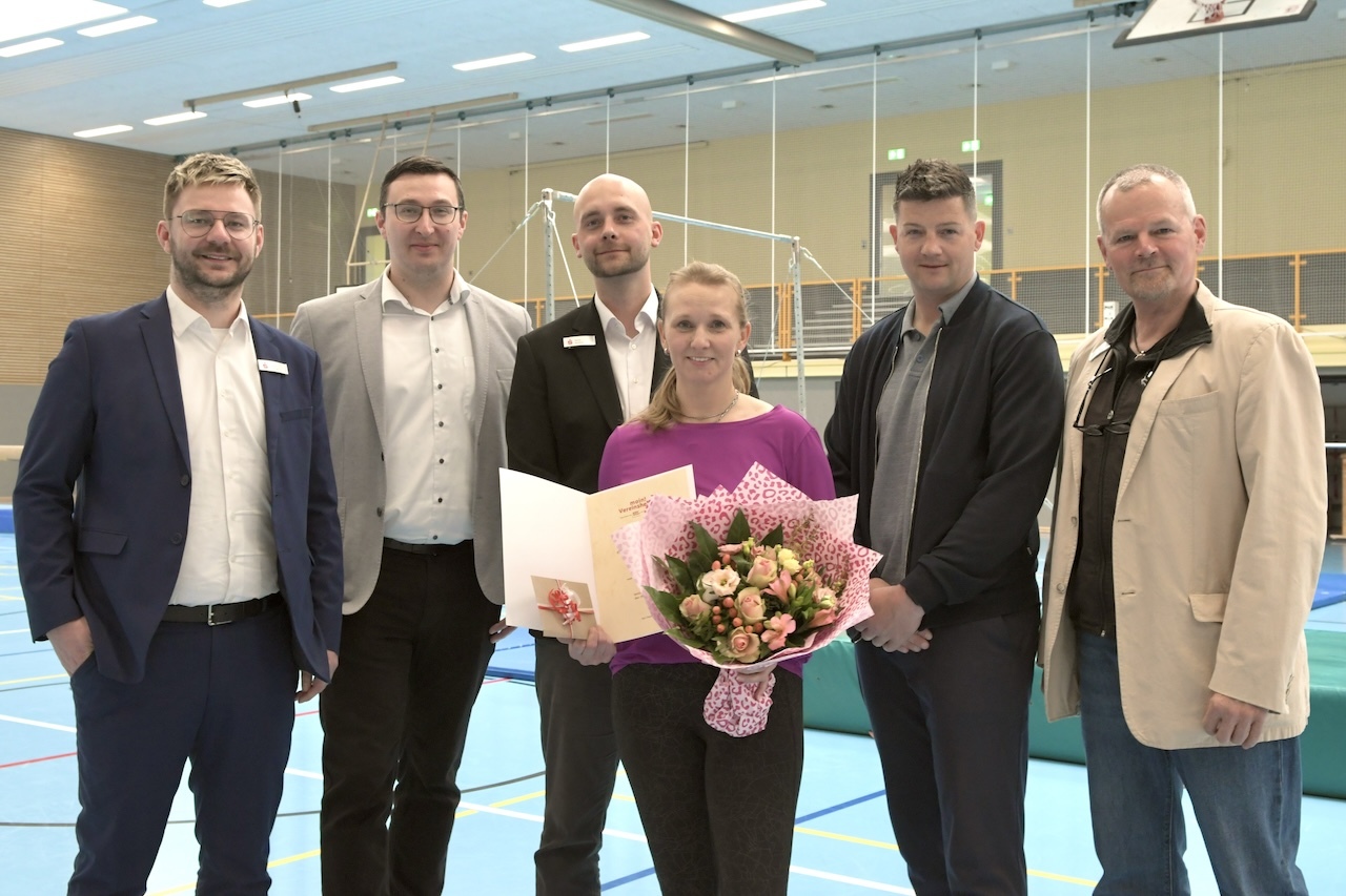 Sechsk&ouml;pfige Gruppe von Menschen in Sporthalle, die eine Frau mit Blumenstrau&szlig; und Urkunde feiert.