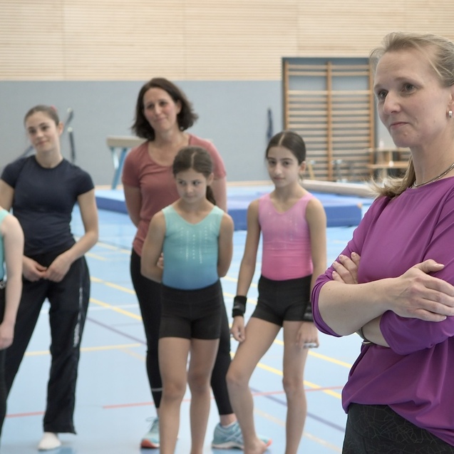 Gruppe junger Turnerinnen und Trainerinnen in einer Sporthalle, eine Trainerin beobachtet aufmerksam das Geschehen.