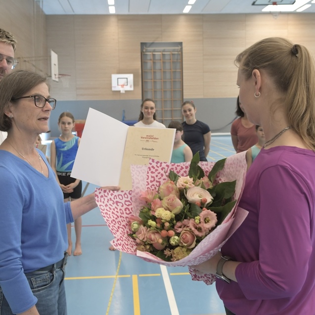 Frau &uuml;berreicht einer anderen Frau einen Blumenstrau&szlig; und ein Dokument in einer Sporthalle, im Hintergrund Sportlerinnen.