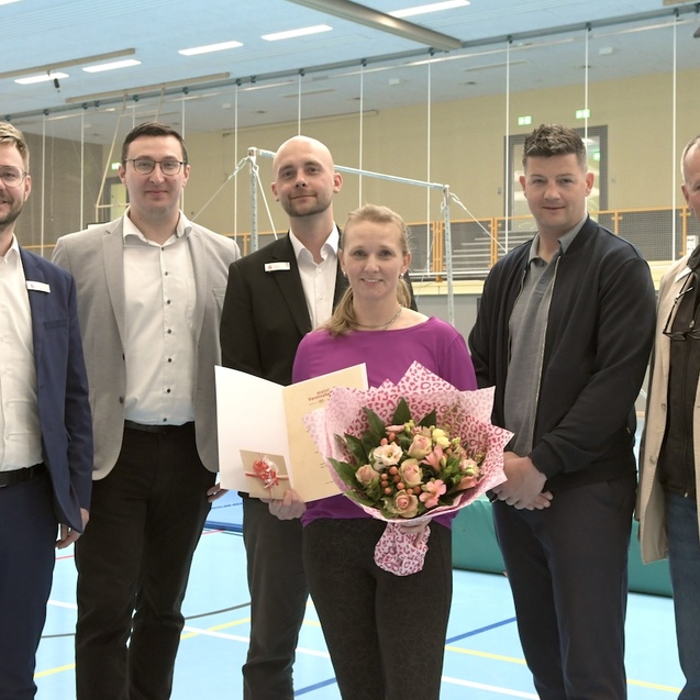 Sechsk&ouml;pfige Gruppe von Menschen in Sporthalle, die eine Frau mit Blumenstrau&szlig; und Urkunde feiert.