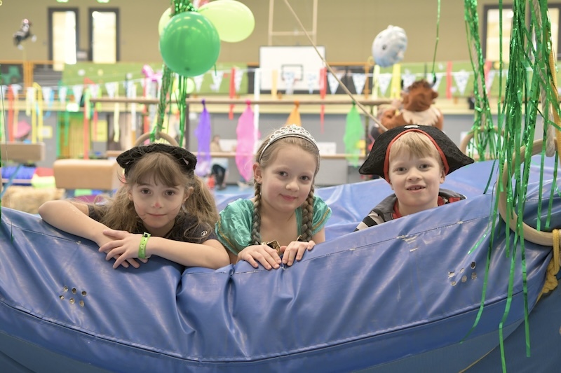 Drei Kinder in Kost&uuml;men lehnen an einer blauen Spielunterlage in einer bunten Indoor-Spielumgebung.