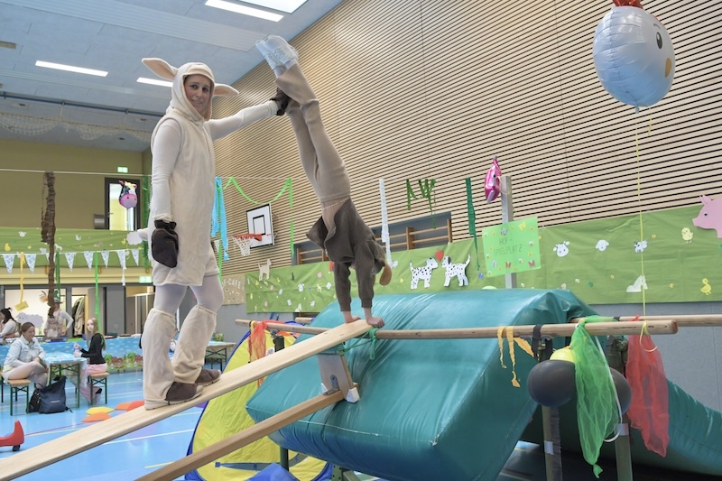 Personen in Tierkost&uuml;men balancieren auf einem Holzbrett &uuml;ber einer bunten Gymnastikunterlage in einer Sporthalle.