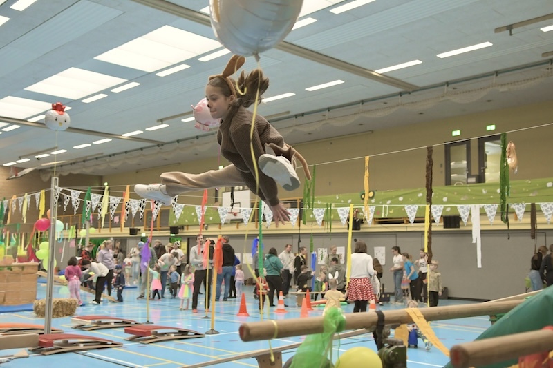M&auml;dchen in Tierkost&uuml;m schwingt mit Luftballon in einer bunten Turnhalle, umgeben von Kindern und festlicher Dekoration.