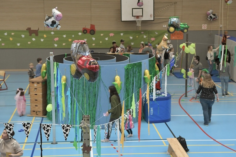 Bunte Festdekoration in einer Sporthalle mit Spielst&auml;nden und Kindern in Kost&uuml;men. Ballons und Girlanden zieren den Raum.