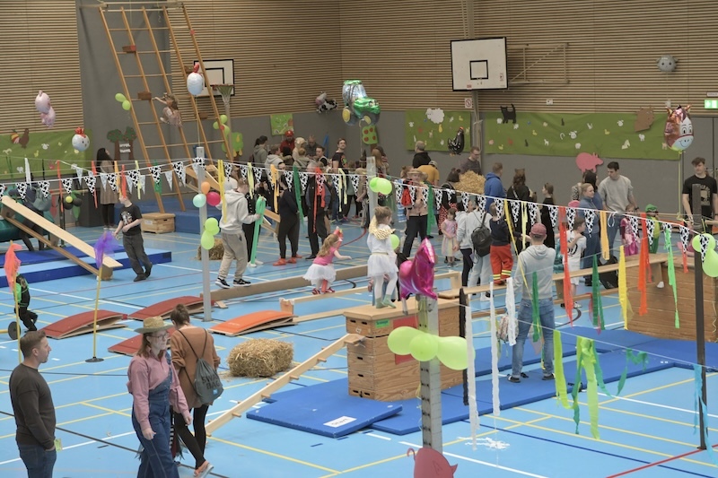 Bunt geschm&uuml;ckte Turnhalle mit Kindern und Erwachsenen, die &uuml;ber H&uuml;pfger&auml;te und Balance-Elemente spielen.