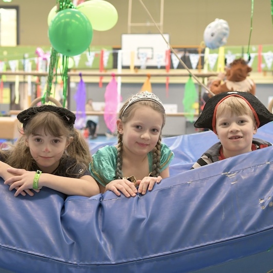 Drei Kinder in Kost&uuml;men lehnen an einer blauen Spielunterlage in einer bunten Indoor-Spielumgebung.