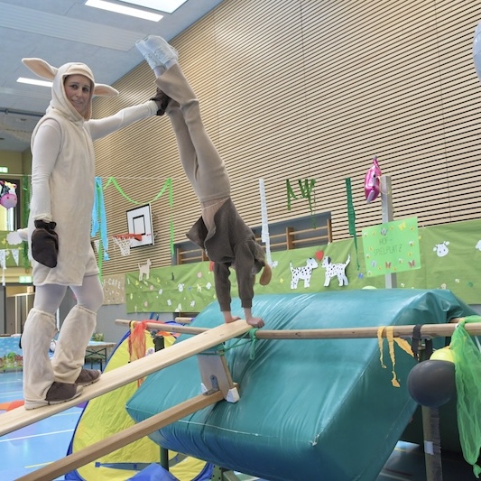 Personen in Tierkost&uuml;men balancieren auf einem Holzbrett &uuml;ber einer bunten Gymnastikunterlage in einer Sporthalle.