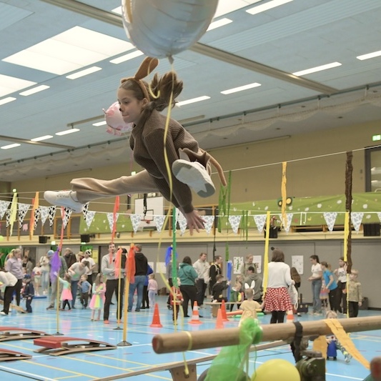 M&auml;dchen in Tierkost&uuml;m schwingt mit Luftballon in einer bunten Turnhalle, umgeben von Kindern und festlicher Dekoration.