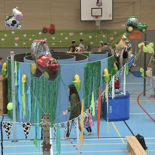 Bunte Festdekoration in einer Sporthalle mit Spielst&auml;nden und Kindern in Kost&uuml;men. Ballons und Girlanden zieren den Raum.