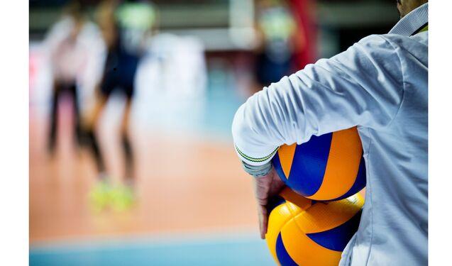 Nahaufnahme einer Person mit zwei Volleyballs in der Hand, im Hintergrund ist ein Volleyballspiel zu sehen.
