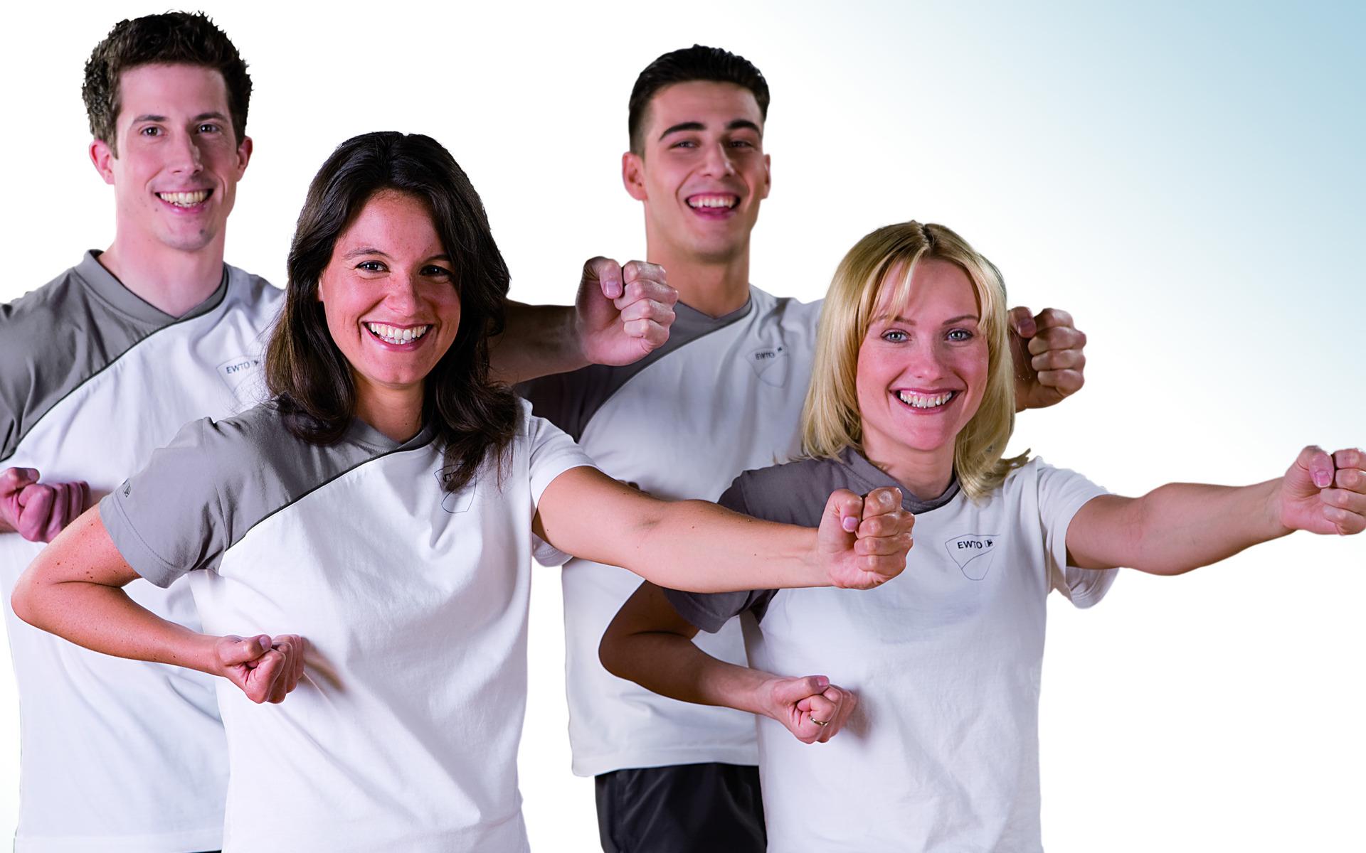 Vier sportliche Personen in hellen T-Shirts posieren freudig mit erhobenen Armen vor einem hellen Hintergrund.