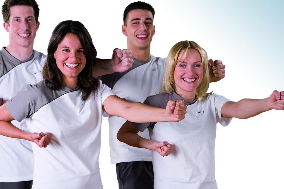 Vier sportliche Personen in hellen T-Shirts posieren freudig mit erhobenen Armen vor einem hellen Hintergrund.