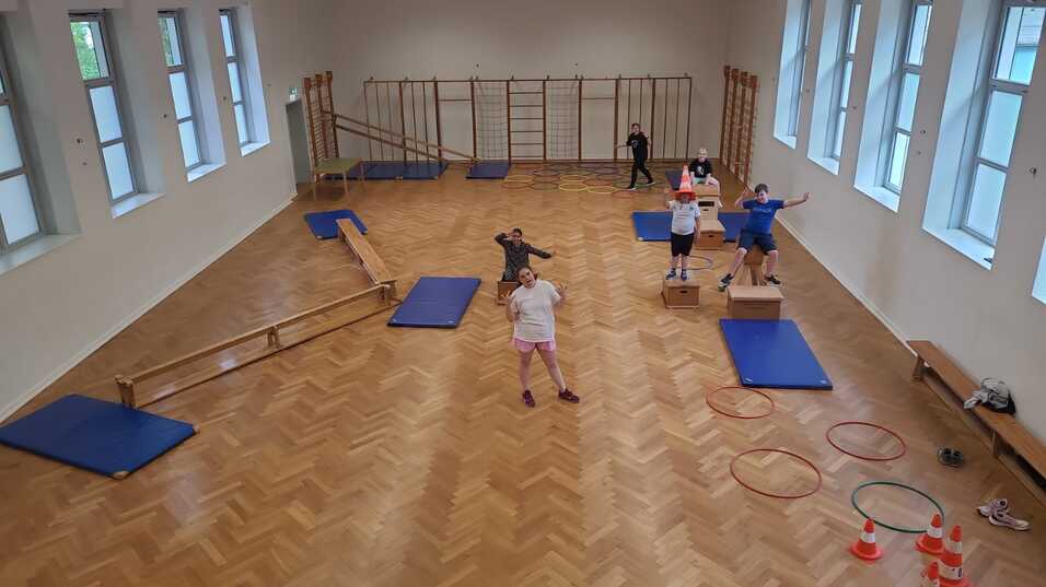 Kinder trainieren in einer Sporthalle mit Matten, Bänken und Geräten zur Förderung der motorischen Fähigkeiten.