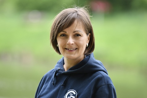 Frau mit schulterlangen, braunen Haaren und blauer Kapuzenjacke l&auml;chelt vor einer gr&uuml;nen Wiese neben einem Gew&auml;sser.